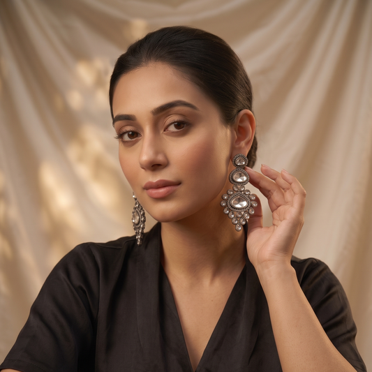A woman in a black V-neck top poses with ornate silver-toned statement earrings while touching one earring. She is seated against a draped beige fabric backdrop, with a gold hand mirror on a black surface in the foreground.