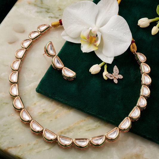 Gold-toned kani necklace with teardrop-shaped white stones, displayed on a dark green velvet cloth over a white marble slab, with a white orchid flower and matching earrings nearby.