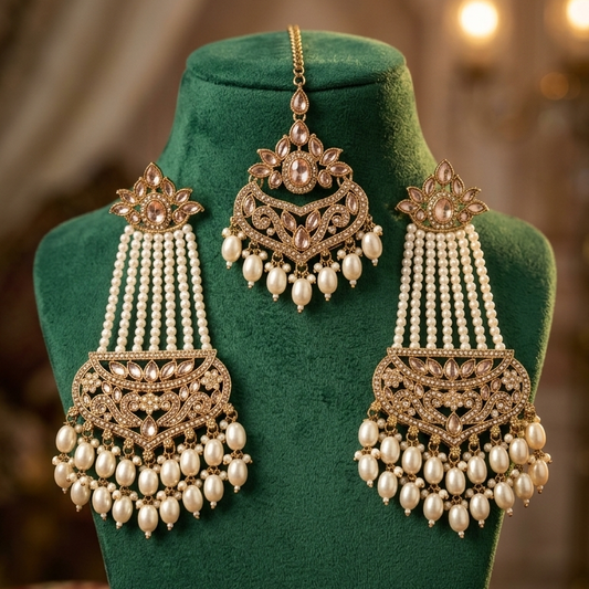 An ornate Indian bridal jewelry set featuring a maang tikka and matching earrings displayed on a green velvet jewelry stand. The jewelry is made of gold-toned metal with intricate filigree work, adorned with white pearls and small clear stones, set against a warm, softly blurred background.