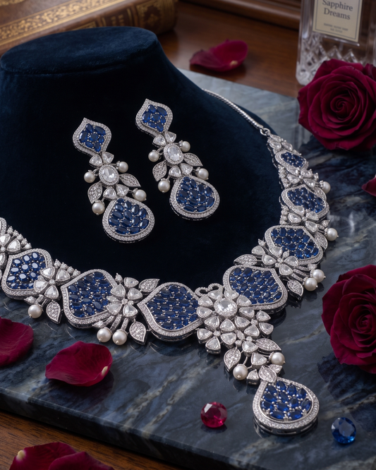 An ornate jewelry set displayed on a dark velvet stand, including a necklace and matching earrings with large blue stones and silver-toned floral detailing, set on a marble slab with rose petals and red roses.