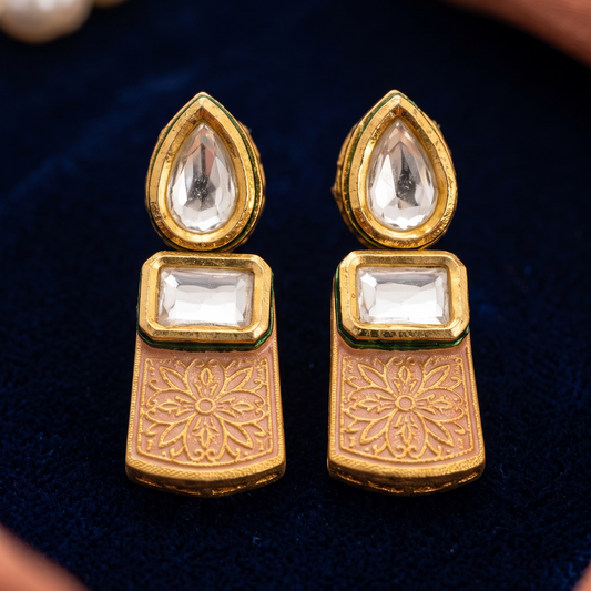 A pair of gold-plated Indian Kundan earrings with clear teardrop and rectangular stones, displayed on a dark blue velvet surface with decorative props.