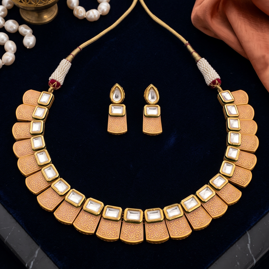 Gold-toned statement necklace and matching drop earrings displayed on a dark blue velvet surface, styled with a brass vessel, white pearls, and peach fabric.
