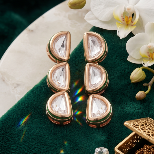 A pair of gold-toned teardrop-shaped earrings with clear stones, arranged on a dark green velvet surface with white orchids, loose stones, and a gold jewelry box nearby.