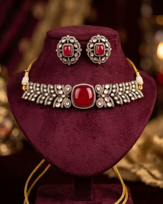 A traditional Indian jewelry set displayed on a dark burgundy velvet stand, featuring a gold-plated necklace with a large red cabochon stone and matching stud earrings, set against a dark, blurred background.