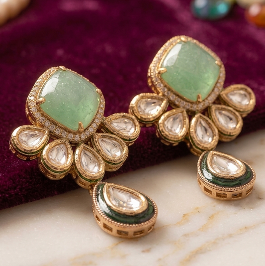 A pair of traditional Indian gold-plated earrings with green stones and clear crystals, displayed on a white marble slab over a deep purple velvet fabric.