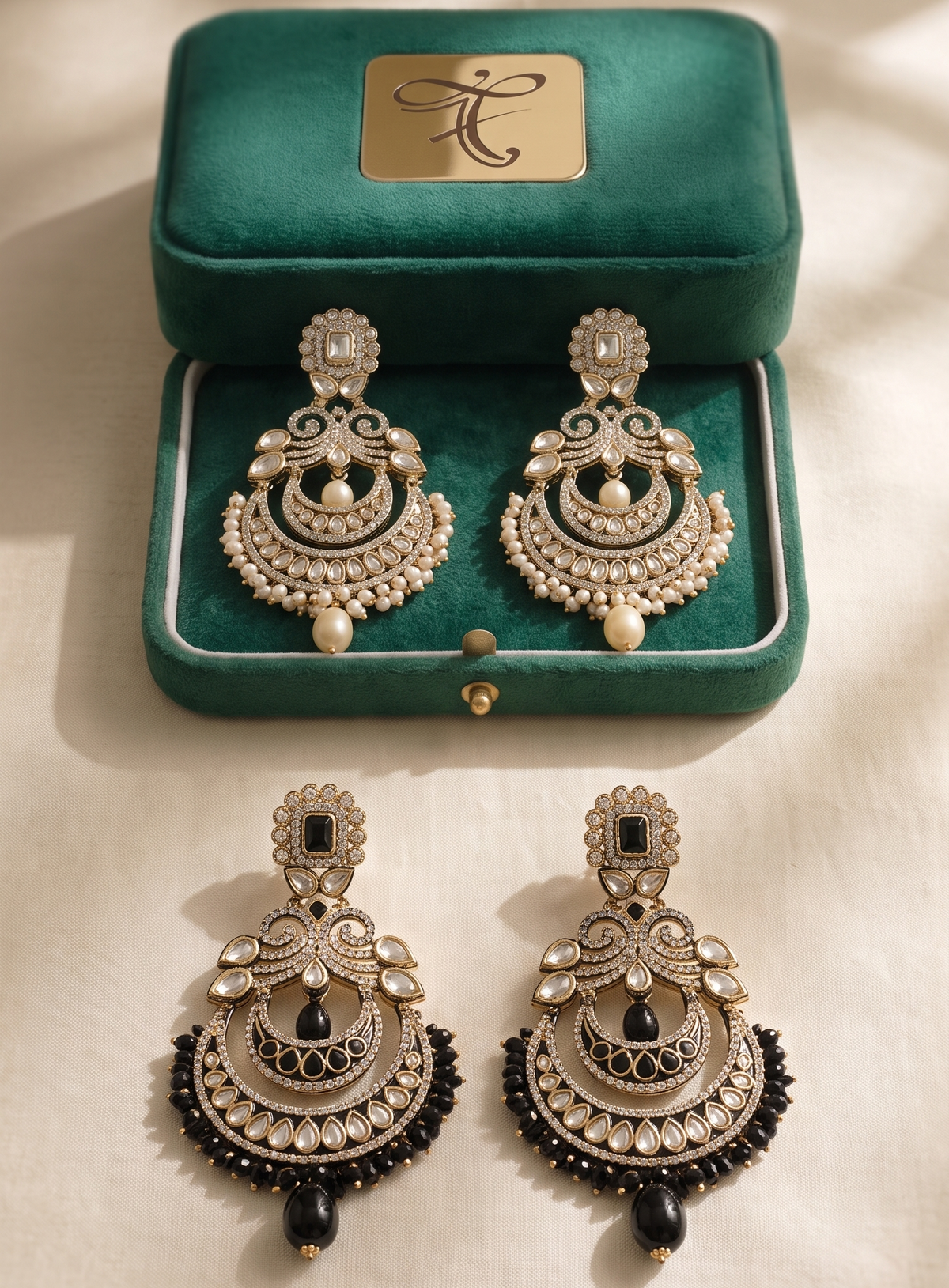 A pair of ornate gold-toned chandelier earrings with black bead accents and white pearl-like details, displayed below an open green jewelry box on a cream fabric surface with soft shadows.