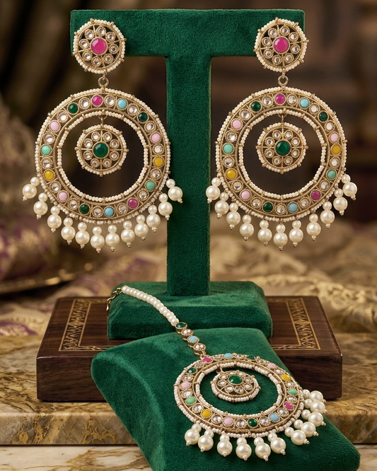 An ornate Indian jhumka earrings and matching pendant set displayed on a green velvet stand. The jewelry features intricate gold-plated filigree, circular motifs, and is embellished with pink, green, yellow, and blue stones, finished with dangling white pearl chains.