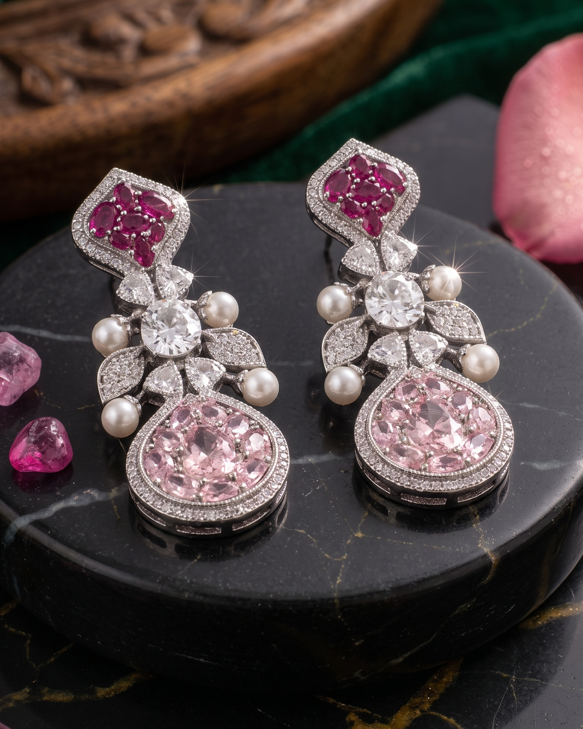 A pair of ornate dangle earrings displayed on a black marble surface. Each earring features a silver-toned setting with a large round pink gemstone at the bottom, a cluster of clear stones and pearls in the middle, and a smaller pink gemstone at the top.