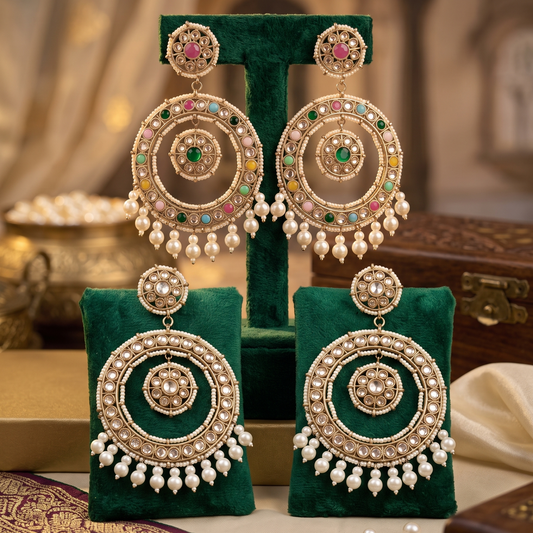 A pair of ornate gold-toned dangle earrings with multicolored stone accents and pearl bead drops, displayed on green velvet jewelry stands against a warm, softly blurred background.