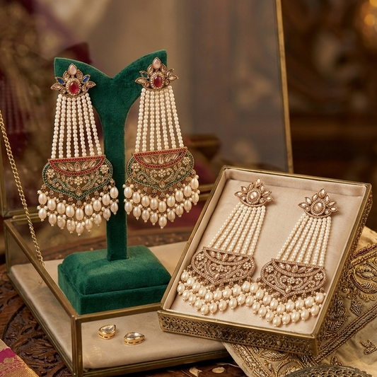 A pair of ornate gold and pearl jhumka earrings displayed on a green velvet stand next to their matching box, with a small mirror and decorative accessories on a carved wooden table.