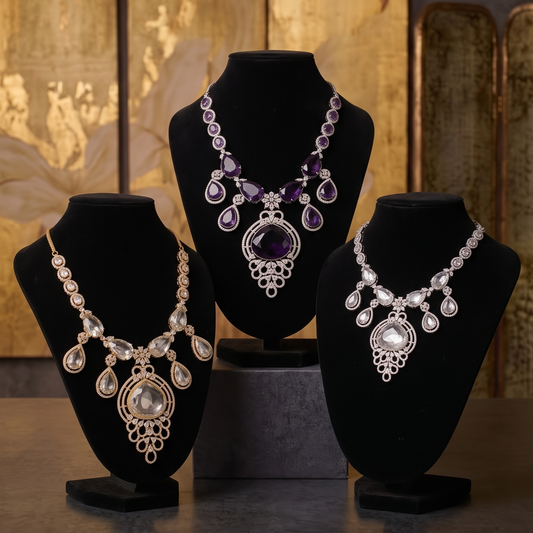 Three ornate statement necklaces displayed on black jewelry busts against a warm gold-toned backdrop. The center necklace is silver with large purple teardrop stones and a purple central pendant.
