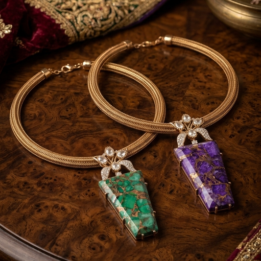 A pair of gold-toned traditional Indian earrings with rectangular stone pendants, one green and one purple, displayed on a wooden surface with traditional Indian textiles and a brass bowl in the background.