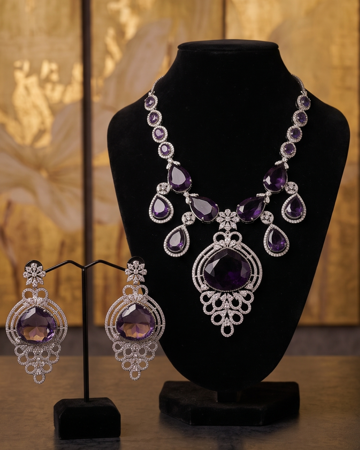 A luxury jewelry set featuring a silver-toned necklace and matching drop earrings with large oval purple amethyst stones, displayed on black velvet stands against a warm, golden-toned background.