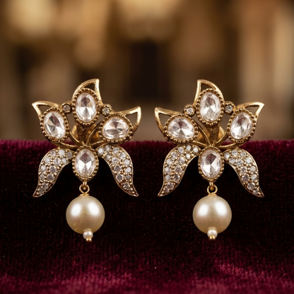A pair of gold-toned floral earrings with clear rhinestone accents and dangling pearl drops, displayed on a deep burgundy velvet surface against a softly blurred warm brown background.