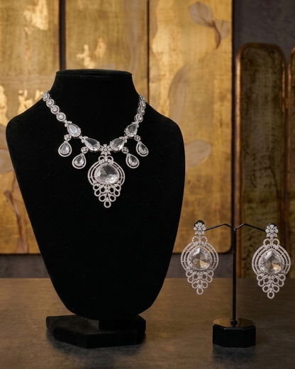 A silver-toned statement necklace displayed on a black velvet bust, with matching drop earrings on a black stand beside it, set against a dark background with gold-toned decorative panels.