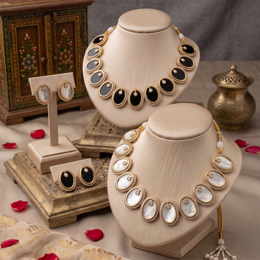 Two cream-colored jewelry busts display ornate gold-tone necklaces with oval white shell-like centers and black inlays, alongside matching stud earrings on a styled tabletop with red petals, a small brass vessel, and a decorative cabinet.
