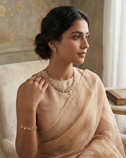 A woman wearing a light beige saree and matching jewelry sits on a cream-colored armchair in a softly lit room with patterned wallpaper.