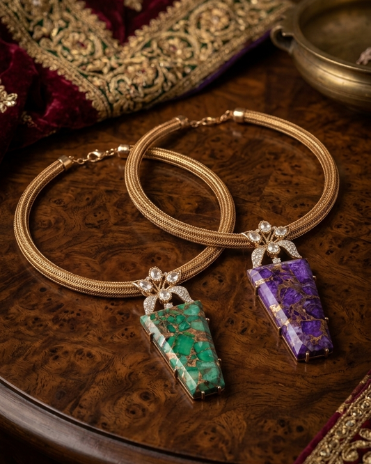A pair of gold-toned traditional Indian earrings with rectangular stone pendants, one green and one purple, displayed on a wooden surface with traditional Indian textiles and a brass bowl in the background.