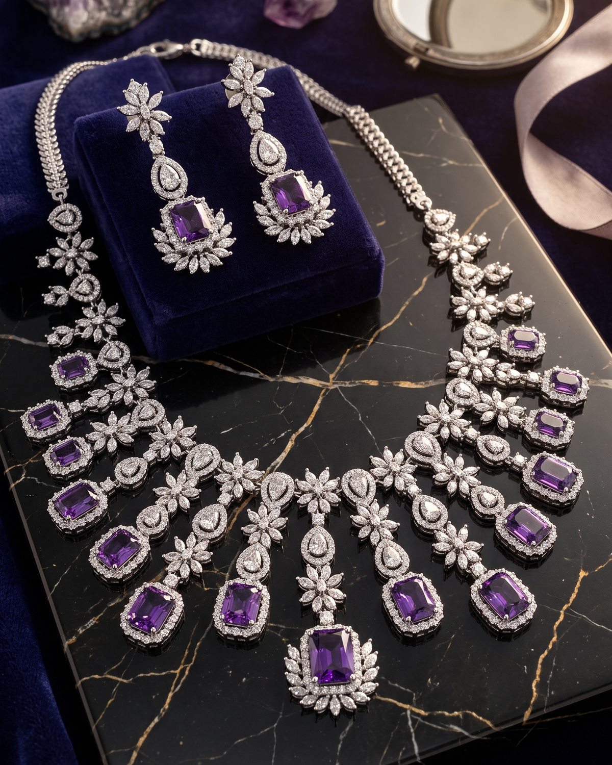 An ornate silver-toned necklace and matching drop earrings with large purple gemstones and clear stones, displayed on a dark velvet jewelry box. The jewelry is arranged on a black marble slab with purple crystals and a small round mirror in the background.