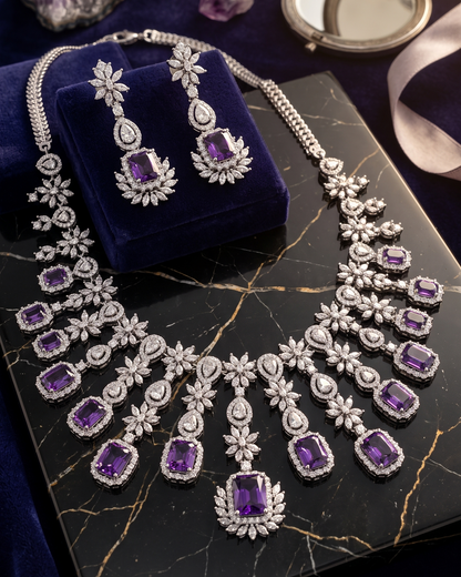 An ornate silver-toned necklace and matching drop earrings with large purple gemstones and clear stones, displayed on a dark velvet jewelry box. The jewelry is arranged on a black marble slab with purple crystals and a small round mirror in the background.