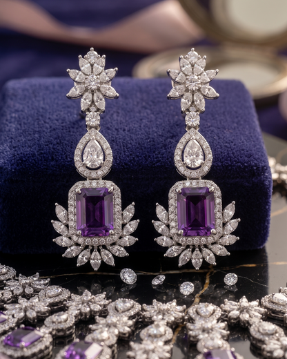 A pair of luxury drop earrings with large emerald-cut purple gemstones and surrounding pavé-set clear stones, displayed on a dark blue velvet jewelry box over a black marble surface with other jewelry scattered around.