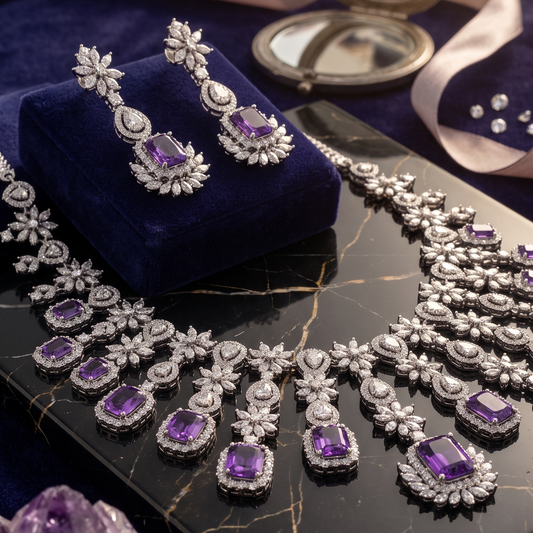 An elegant jewelry set featuring a silver-toned necklace and matching drop earrings with oval purple gemstones, displayed on a dark velvet cushion atop a black marble slab with scattered diamonds, crystals, and a compact mirror.