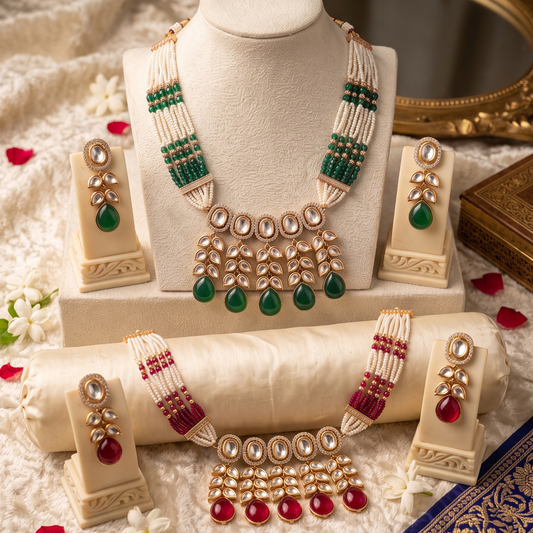 A traditional Indian jewelry set displayed on cream-colored mannequin stands, featuring a necklace, earrings, and a bracelet with white beads, green teardrop stones, and red stones, styled with a gold mirror and fabric background.