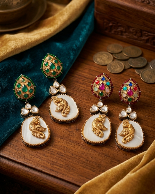 Four ornate gold-toned earrings with green and multicolor gemstone studs, white oval inlays, and raised gold animal motifs, arranged on a wooden surface with teal fabric, gold fabric, and coins as styling props.